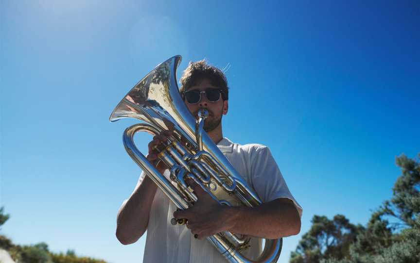 Young man plays the Euphonium with the beach and a bright blue sky in the background