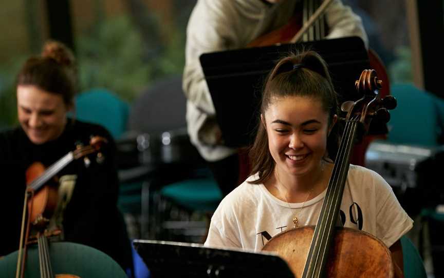 A young person playing cello and smiling with other musicians in the background