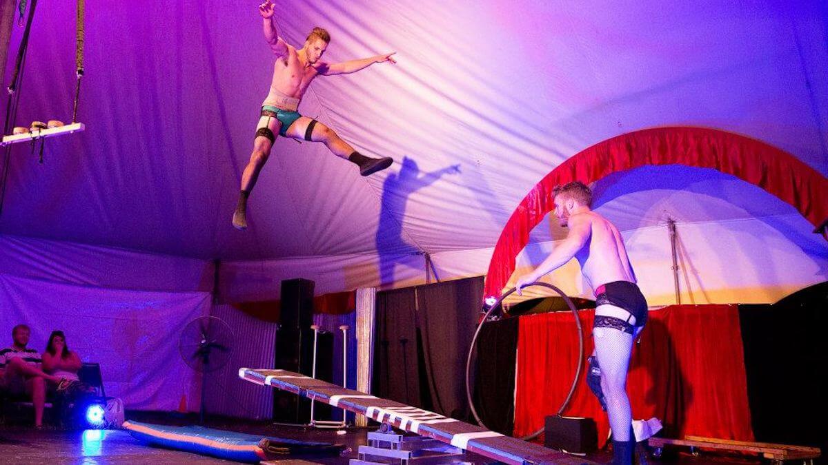two male circus performers in their underwear on stage. One is jumping up high and the other is holding a cyr wheel on the stage
