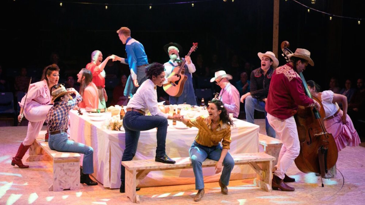 A group of people dressed in American country clothing party around a table on stage, including a band