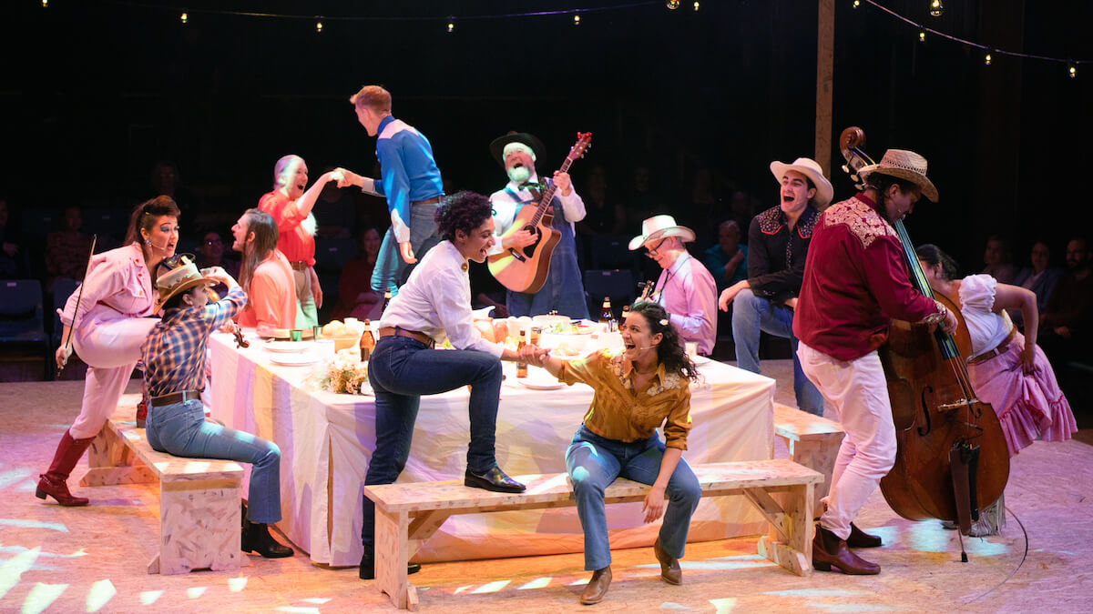 the cast of Oklahoma sitting and standing around a dinner table dressed as cowboys while performing
