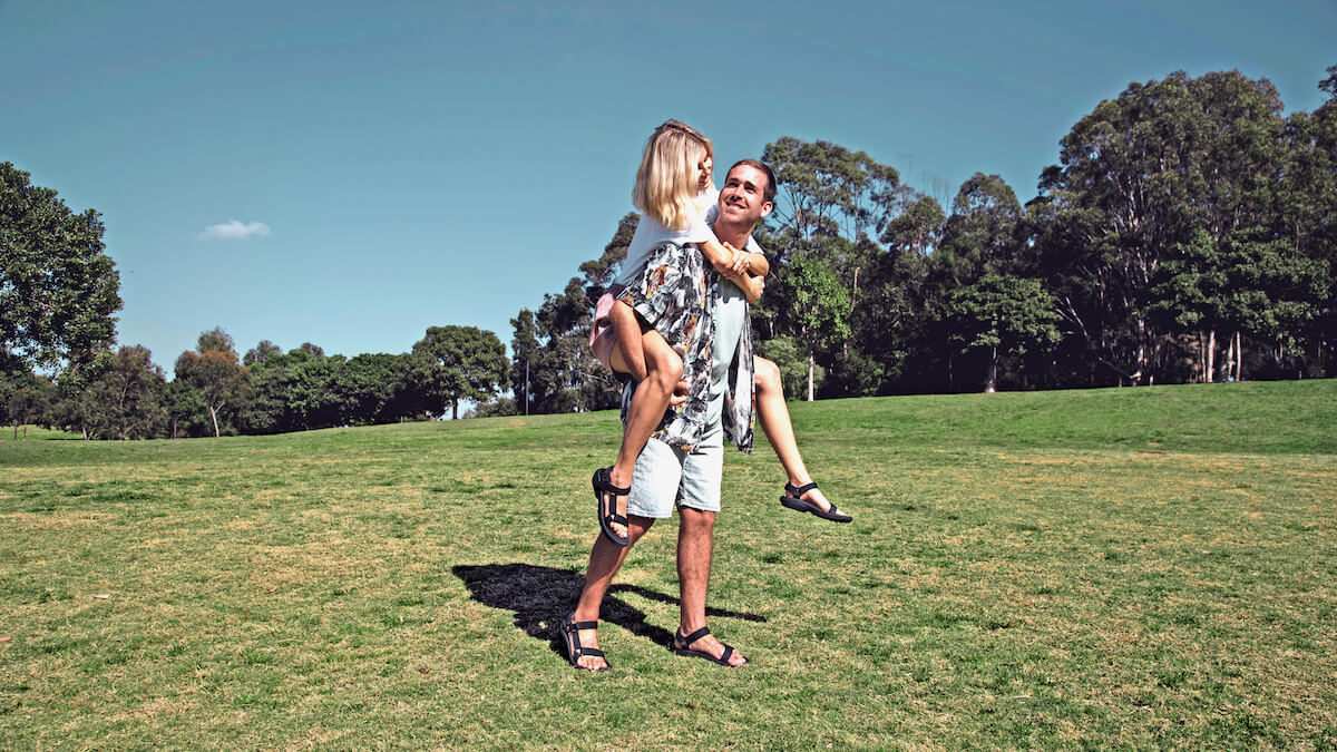 a man dressed for summer wears sandals, and carries a woman, also wearing sandals, on his back. They look relaxed and happy, and are standing in an open grassy space, surrounded by large trees
