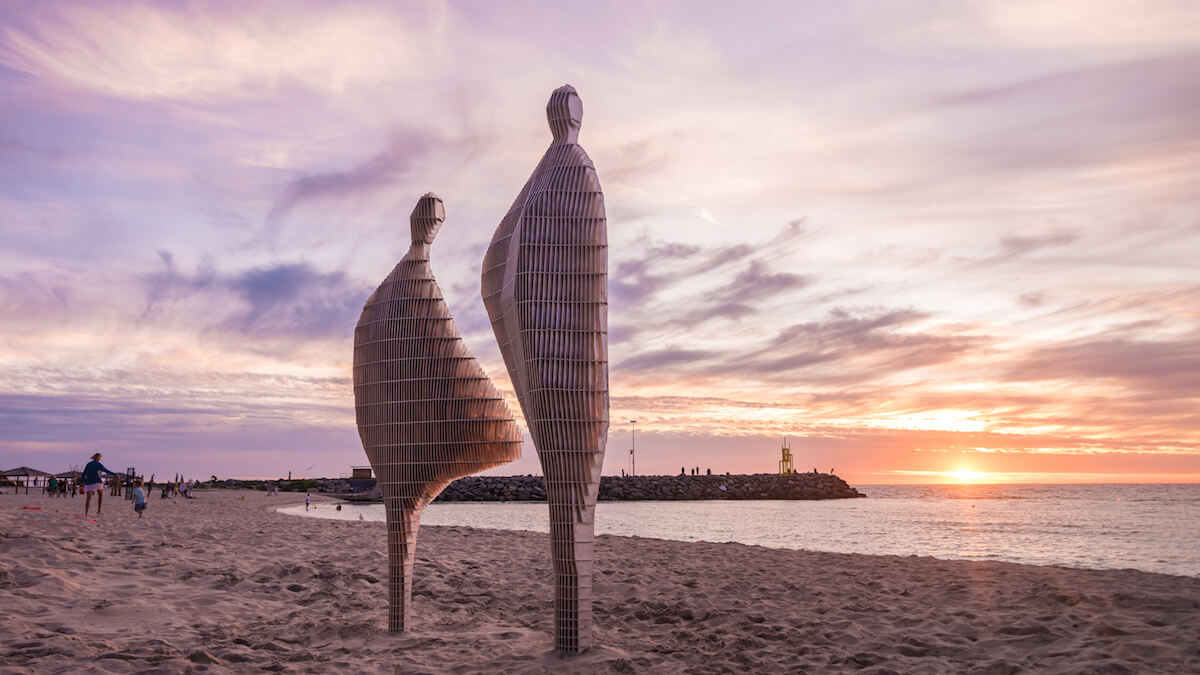 Two abstract scuplted figures installed on the beach in white sand, and appear to be watching the sunset over the water and beach groyne with soft wispy clouds overhead from Sculpture by the Sea 2020