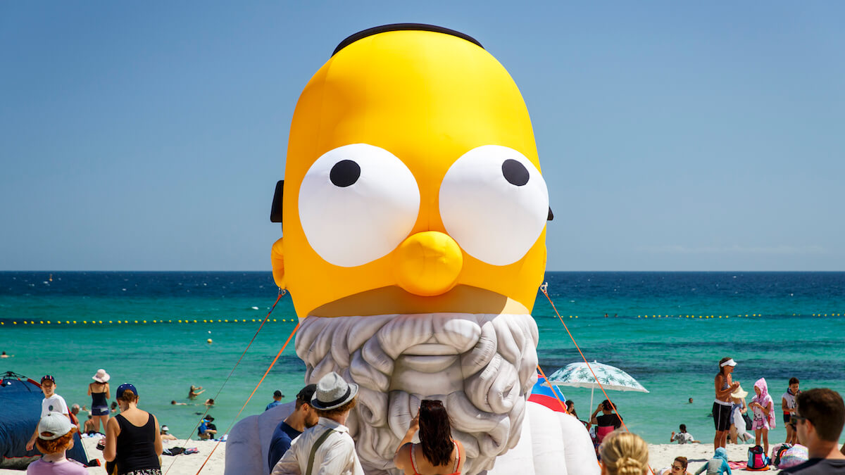 A larger than life inflated head figure sits on the beach, the top half of the head is homer simpson, who has a yellow face and very large round eyes, and the bottom half is the greek poet homer, in the classical sculptural style from Sculpture by the Sea 2020