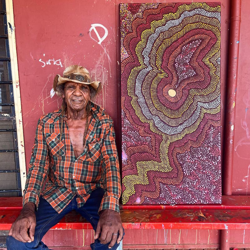 Aboriginal Warlayirti Artist sitting next to his Aboriginal dot painting
