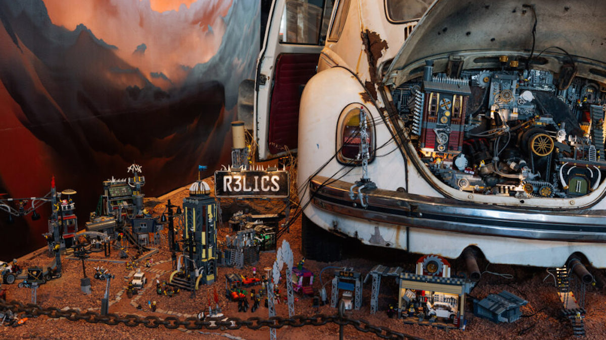 An old, beat-up Volkswagen beetle sits on a bed of dirt. Intricate lego buildings and networks have been built into the boot as well as the ground around below it, resembling a dystopian civilisation and mechanical aspects