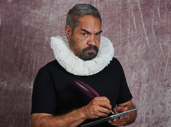 A man wearing a ruff collar and holding a feather quill looks quizically at the camera.
