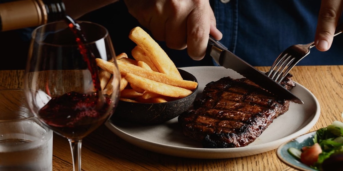 A grilled steak being cut into with chips on the side and a glass of red wine in frame