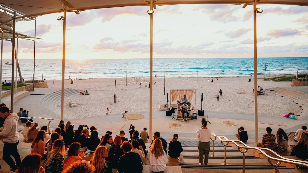 Pictured is the sunset markets at Scarborough beach where a group of people sit in a pavilion facing the beach. 