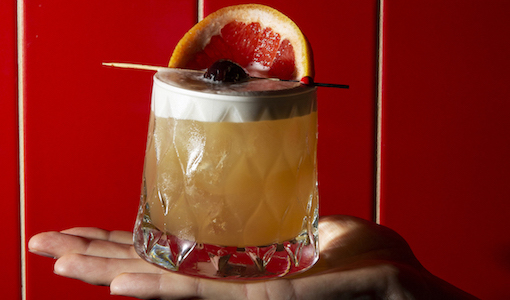 a hand holds an amaretto sour in front of a red tiled wall. There is a blood orange on a toothpick sitting on top of the cocktail